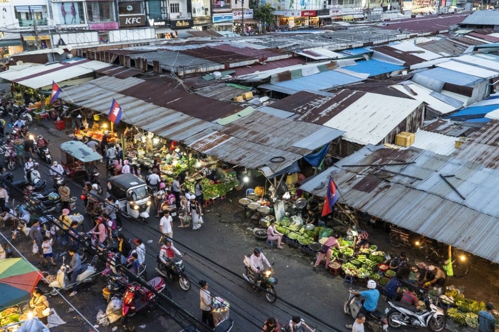 Aerial view of Tuol Tom Pong Market, also known as the Russian Market, featuring bustling vendors, colorful market stalls, and shoppers navigating through narrow walkways. The scene captures the vibrant atmosphere of Phnom Penh's cultural hub, with various goods and fresh produce displayed.