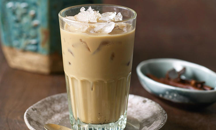 A glass of iced Cambodian coffee with sweetened condensed milk, served over ice, featuring a spoon on a small plate beside it.