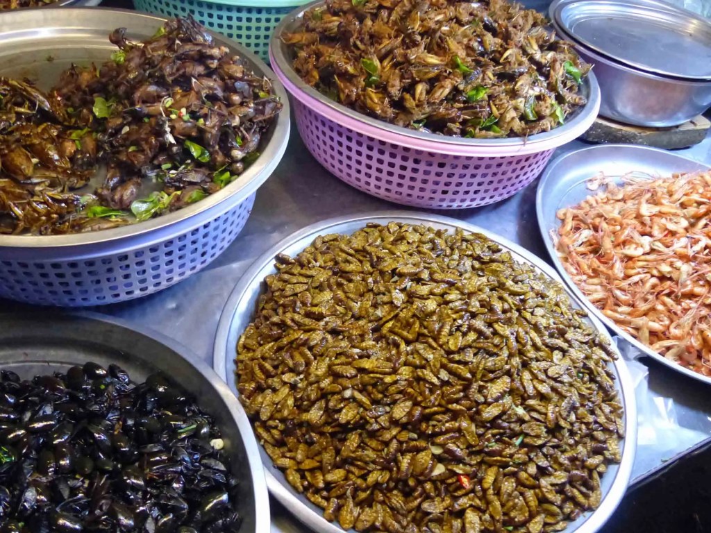 Bowl of fried insects, including crickets and grasshoppers, displayed in colorful baskets at a Cambodian street food market.