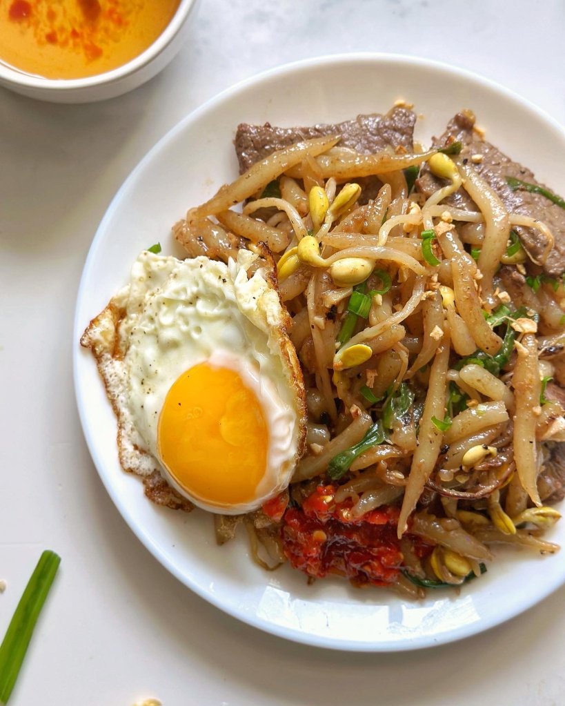 A plate of Cambodian street food featuring stir-fried noodles topped with bean sprouts, slices of beef, a sunny-side-up egg, and a side of chili sauce.
