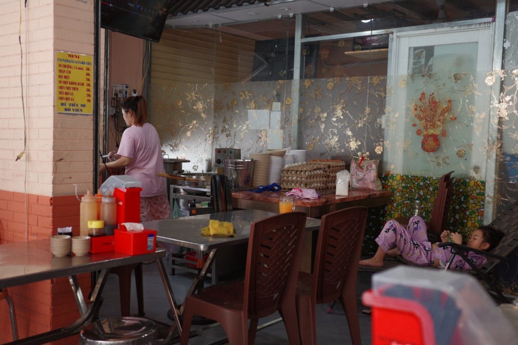 A staff member in a pink shirt preparing food in a small pho restaurant kitchen, with a child relaxing in pajamas nearby.