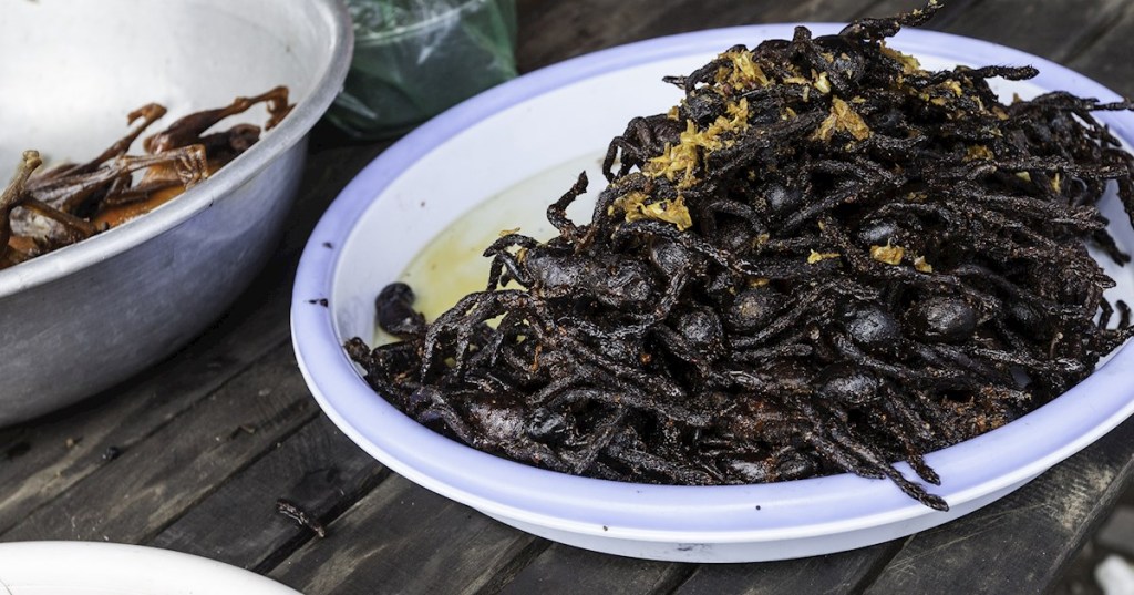 A plate filled with deep-fried tarantulas, served with aromatic seasoning, alongside a bowl of smaller snacks at a street food stall.