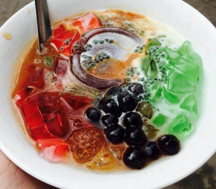 A bowl of Cha Huoy Teuk, a Cambodian dessert featuring colorful jellied agar with coconut milk, palm sugar, and various toppings like mung beans and jackfruit.