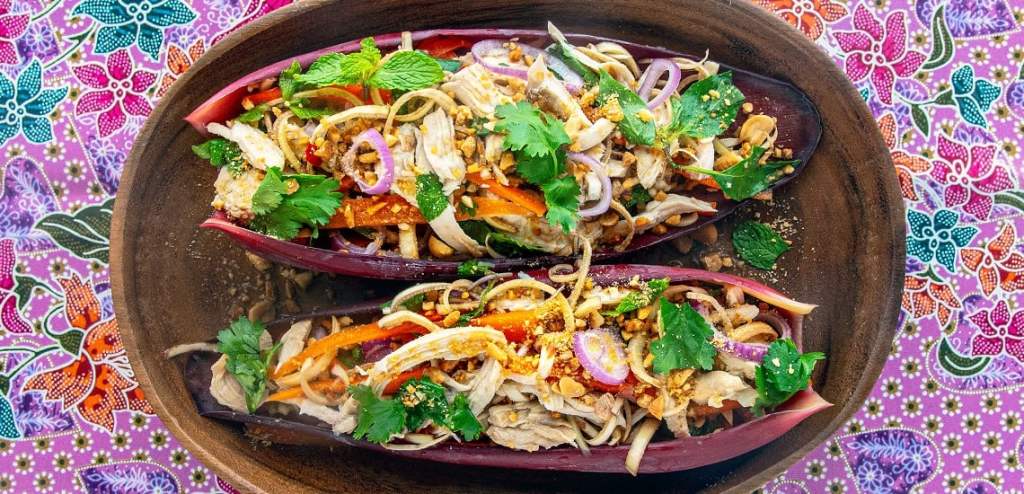 A vibrant Cambodian dish featuring shredded chicken, fresh herbs, and vegetables served in a banana leaf boat, garnished with peanuts and cilantro.