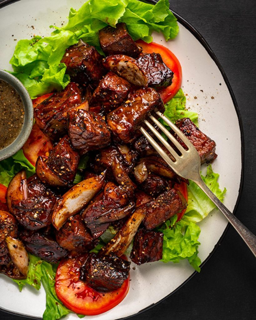 A plate of grilled beef Lok Lak served with lettuce and tomatoes, accompanied by a dipping sauce.