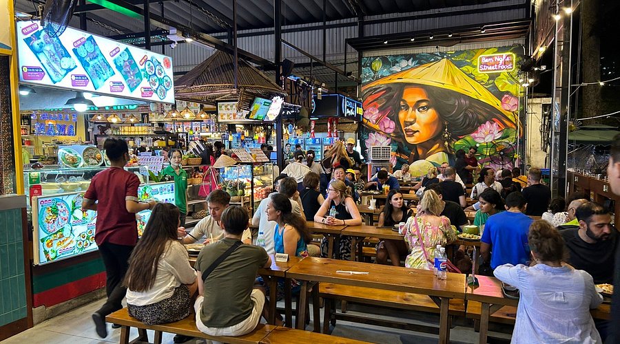 A busy indoor market setting at Bún Nghệ Market in Ho Chi Minh City, showcasing various food stalls with colorful displays, patrons enjoying meals at communal wooden tables, and a vibrant mural in the background.