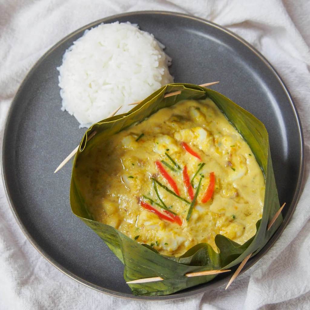 A plate featuring Fish Amok, a traditional Cambodian dish, served in a banana leaf with red chili and green onions, accompanied by a serving of white rice.