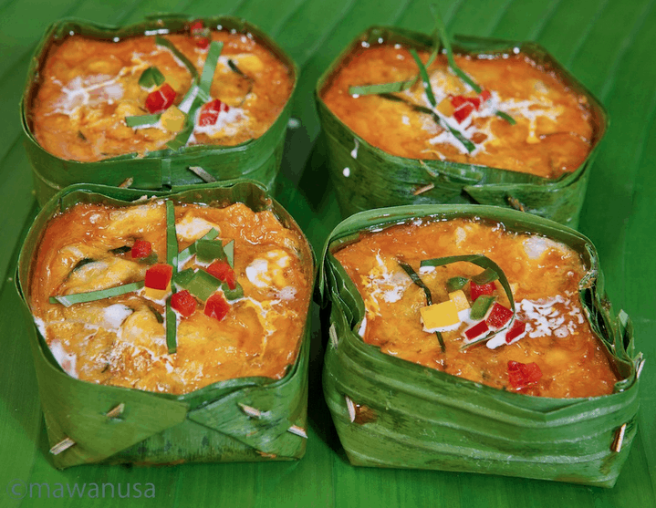Four portions of fish amok, a Cambodian coconut-fish curry, steamed in banana leaves and garnished with red and green peppers, served on a banana leaf.