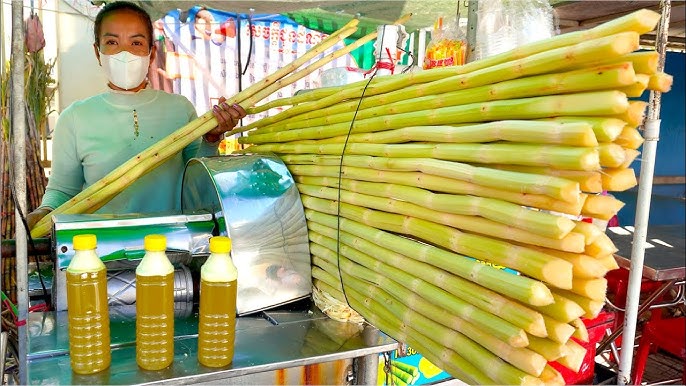 A woman standing behind a cart displaying fresh sugarcane stalks, with bottles of sugarcane juice in front.