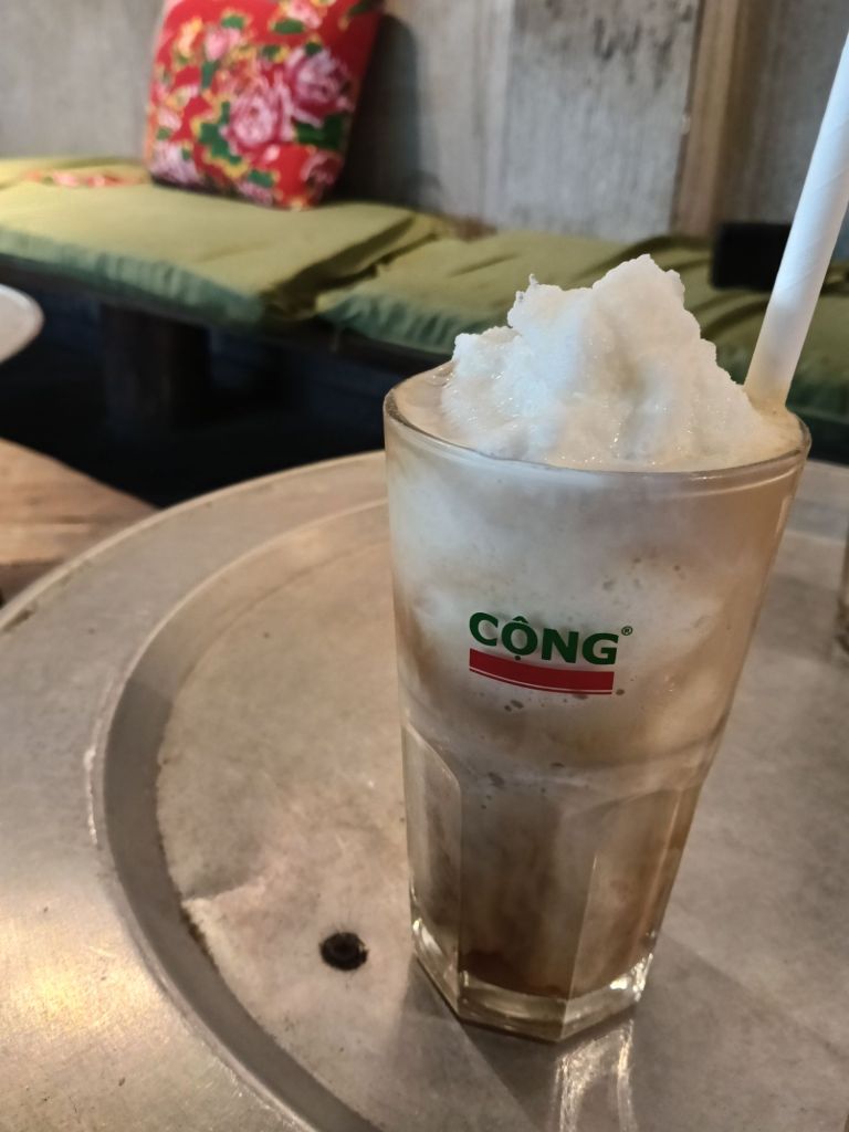A glass of coconut coffee topped with whipped cream, served in a vintage-style cup with the Cộng Cà Phê logo. In the background, colorful cushions and wooden seating create a cozy café atmosphere.