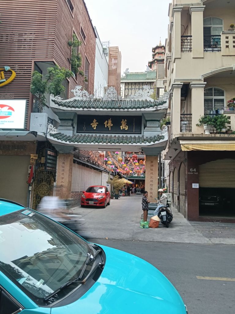 View of the entrance to Ten Thousand Buddha Pagoda in Ho Chi Minh City, showcasing traditional architecture, a decorative archway with Chinese characters, and the bustling street environment.