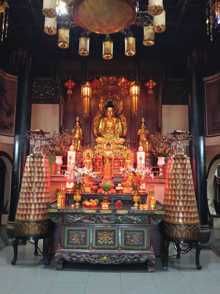 Interior view of the Ten Thousand Buddha Pagoda, featuring a main altar with golden Buddha statues, surrounded by floral offerings and decorative lanterns.
