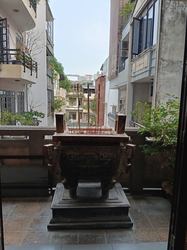 A view from a doorway featuring a traditional incense burner on a pedestal, overlooking a narrow alley with neighboring buildings and greenery.