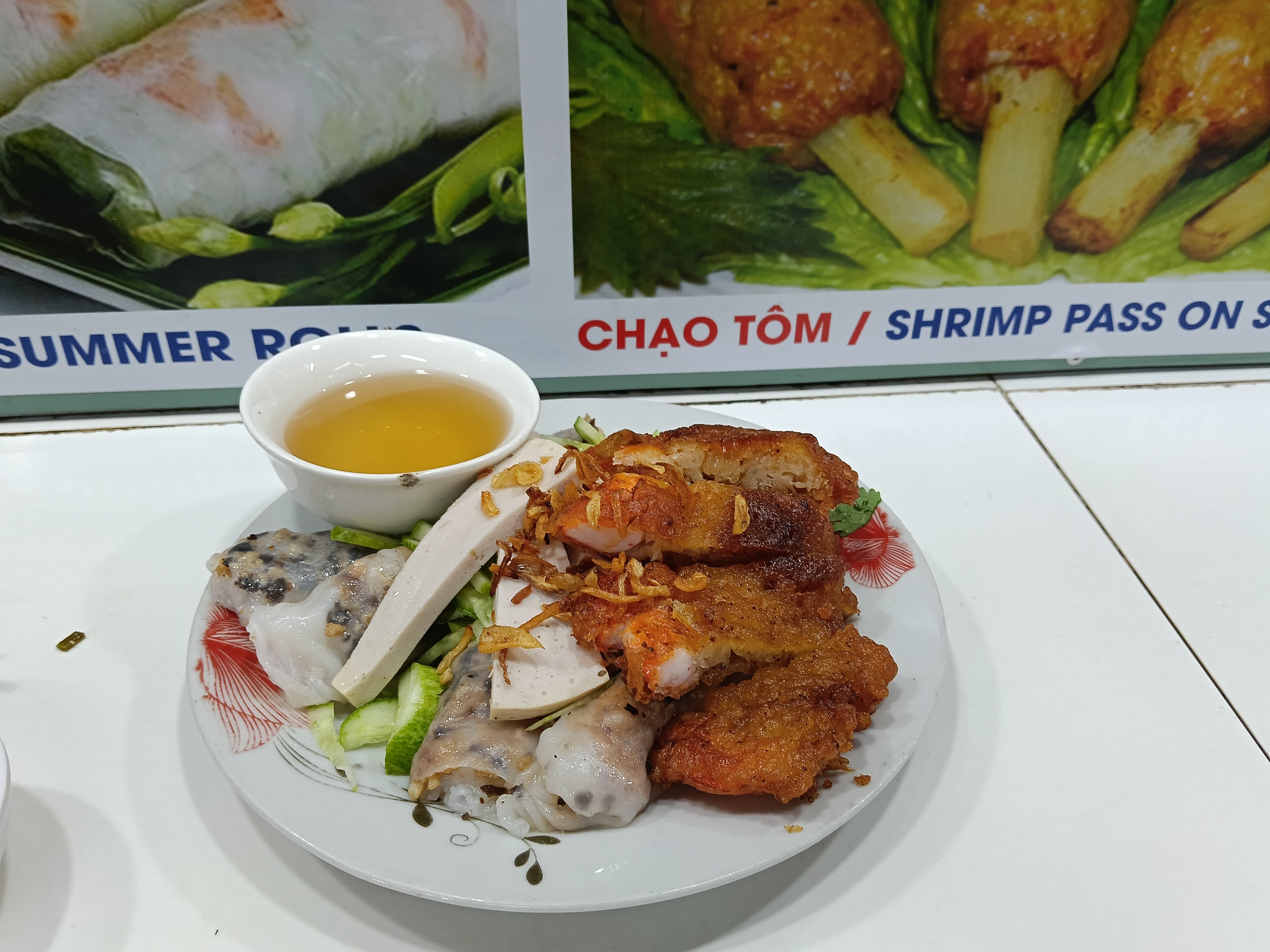 A plate of Vietnamese food featuring shrimp and pork dishes, served with a side of dipping sauce and garnished with herbs, alongside a colorful backdrop of menu items.