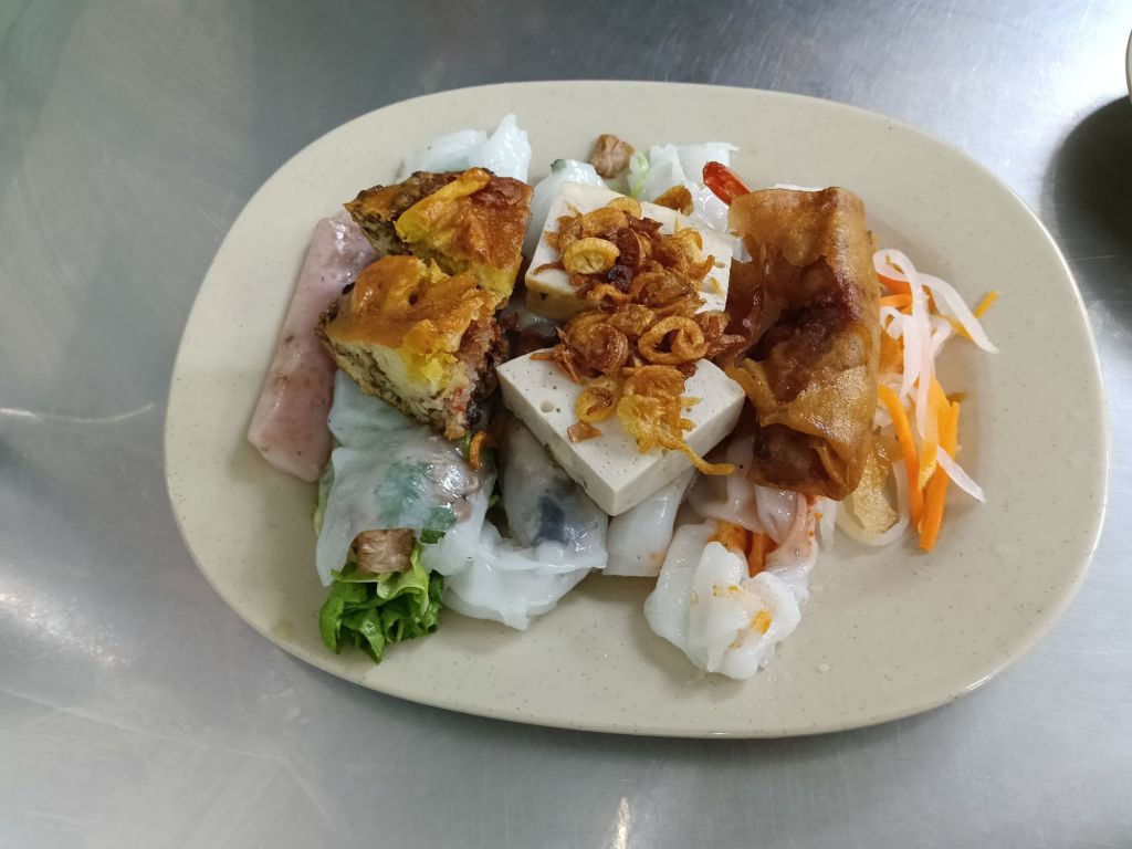 A plate of Bánh Cuốn featuring steamed rice rolls filled with pork and mushrooms, topped with crispy shallots, tofu, and served with pickled vegetables.