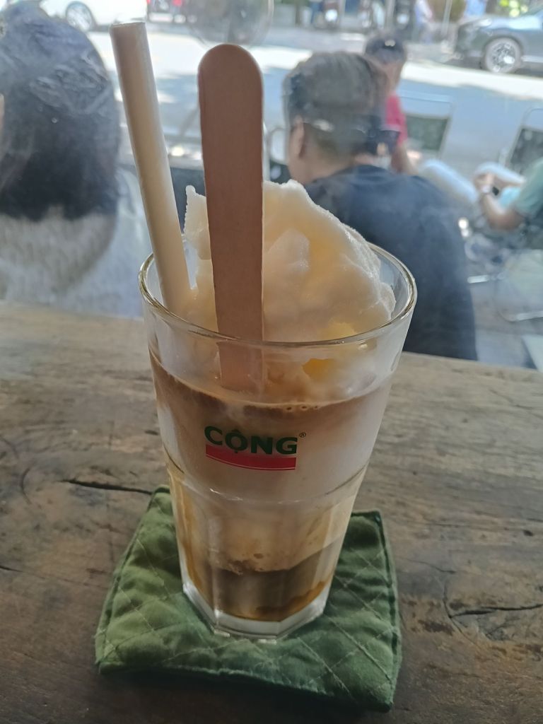 A glass of coconut coffee topped with ice cream, presented with wooden sticks and a straw, placed on a green fabric coaster with a retro-style café ambiance in the background.