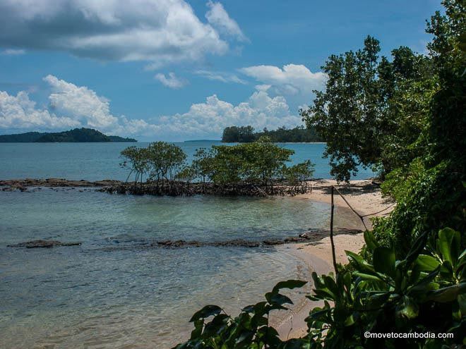 A serene beach scene in Cambodia featuring clear turquoise waters, sandy shores, and lush greenery. The landscape includes rocky formations and distant islands under a bright blue sky with fluffy clouds.