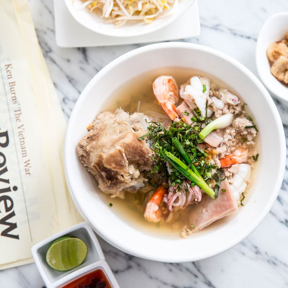 A bowl of kuy teav, a Cambodian noodle soup featuring rice noodles, shrimp, meat, and garnished with green onions, served with lime and dipping sauce.