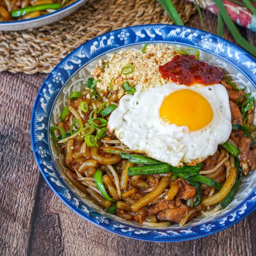 A bowl of stir-fried noodles topped with a sunny-side-up egg, scallions, and chili paste, served on a decorative blue plate.