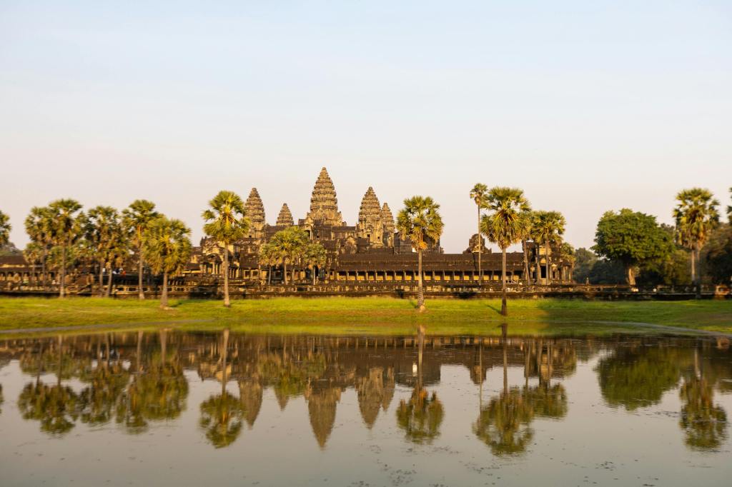 A view of Angkor Wat temple complex in Cambodia, surrounded by palm trees and reflecting in a tranquil body of water, during a soft sunrise.