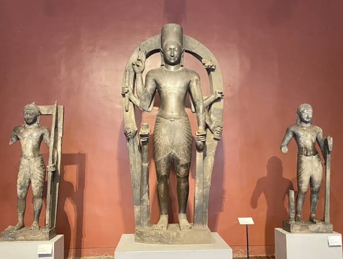 Three ancient Khmer statues displayed in the National Museum of Cambodia, showcasing intricate details and traditional attire against a reddish background.