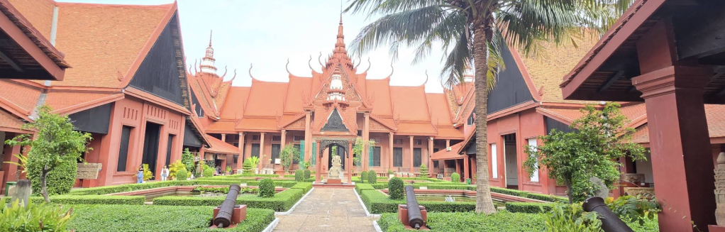 A panoramic view of the National Museum of Cambodia's courtyard, showcasing traditional Khmer architecture with terracotta-red buildings, palm trees, and well-maintained gardens.