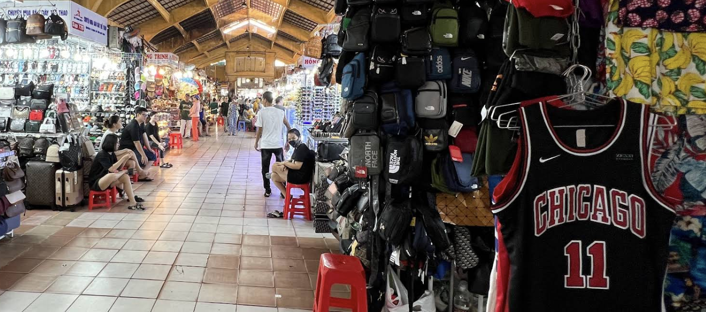 Interior view of Bến Thành Market showing various stalls with bags and clothing, bustling with shoppers and vendors.