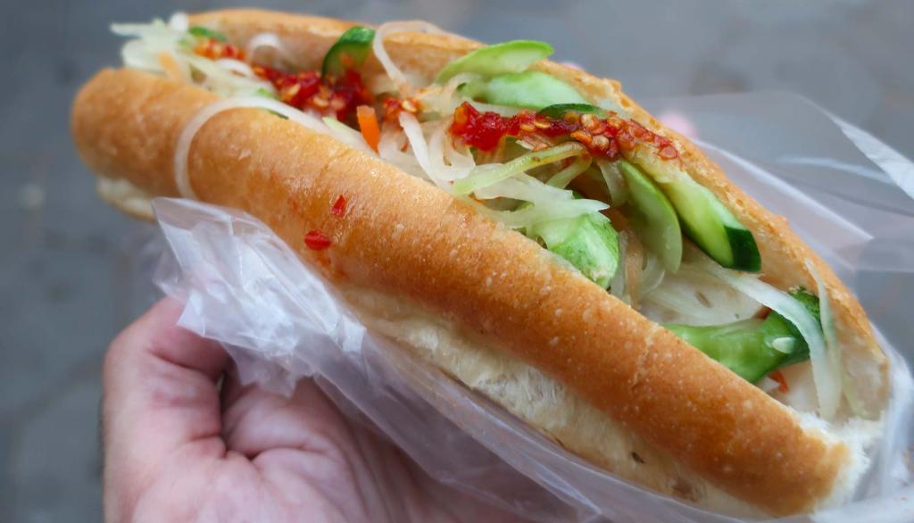 A hand holding a Cambodian Num Pang sandwich filled with vegetables, served in a clear plastic wrapping.