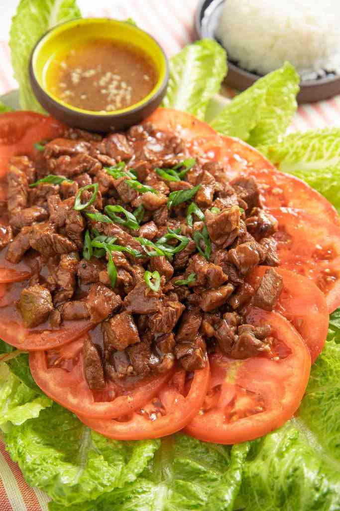 A plate of Lok Lak, a Cambodian dish featuring marinated beef served on a bed of sliced tomatoes and lettuce, accompanied by a small bowl of sauce.