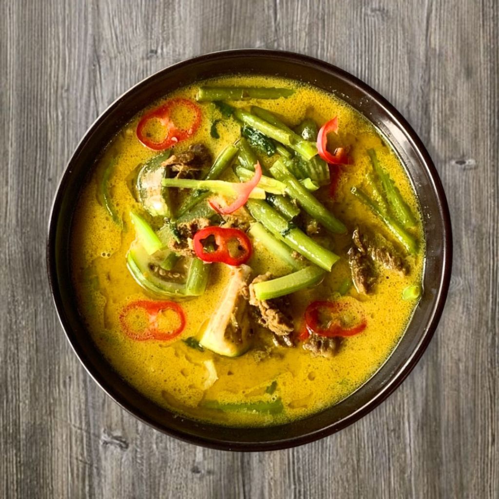 A bowl of Khmer red curry soup featuring yellow coconut broth, chopped green vegetables, and slices of red chili pepper.