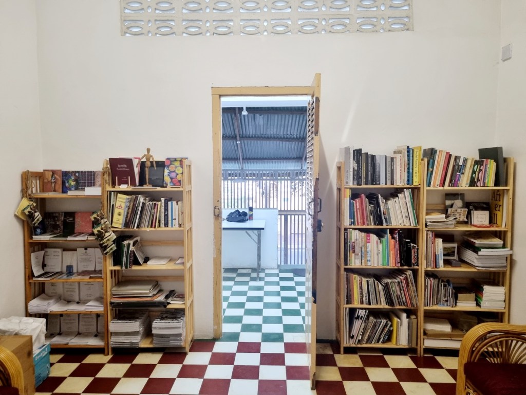 Interior of a minimalist art space featuring wooden bookshelves filled with various art books and materials, with a doorway leading to another room.