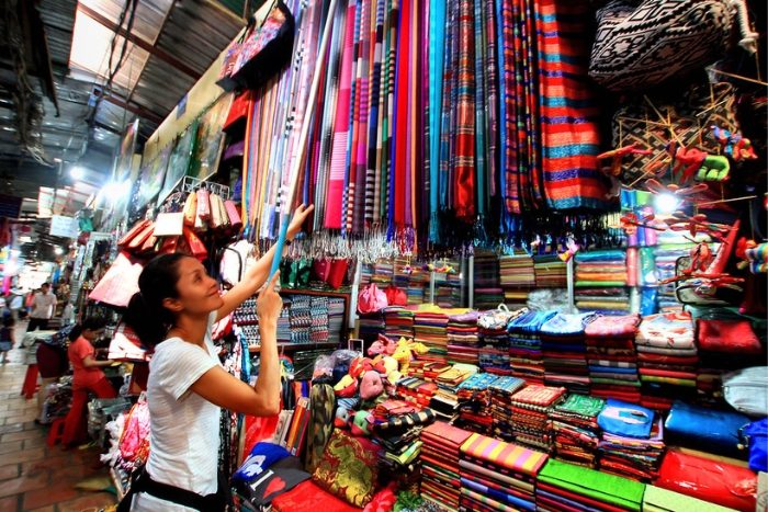 A woman browsing colorful fabrics at a bustling market, surrounded by vibrant textiles and artisan goods.