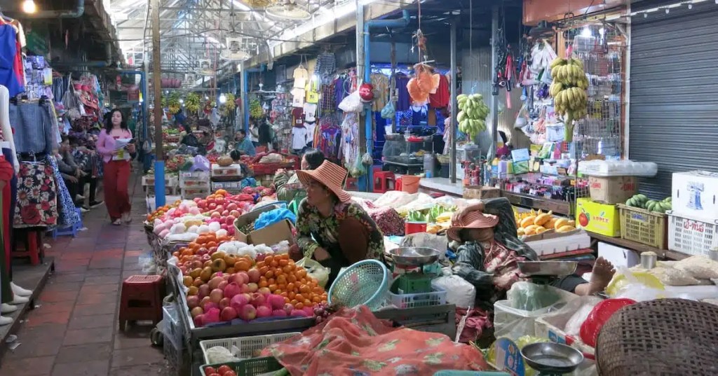 A bustling market scene in Phnom Penh, Cambodia, featuring colorful displays of fruits and vegetables, with vendors and shoppers interacting among stalls selling various goods.