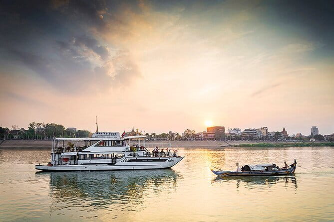 A scenic view of a river during sunset, featuring a large cruise boat and a smaller traditional boat on the water, with the skyline of Phnom Penh in the background.