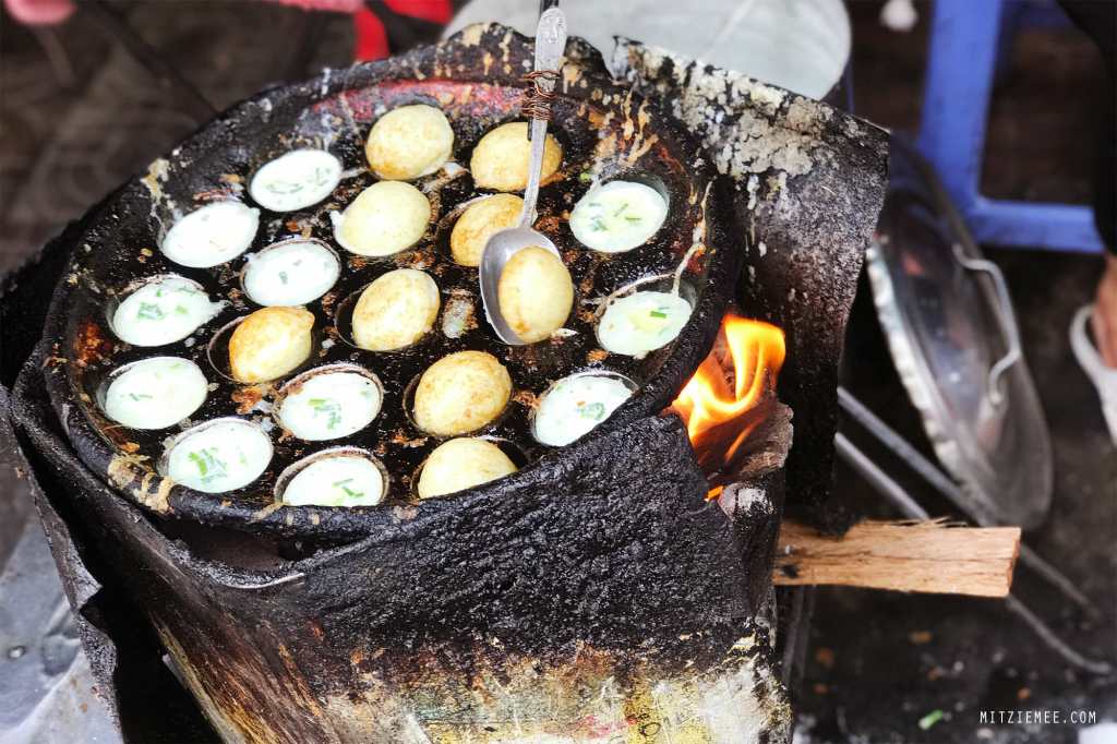 A traditional Cambodian Num Kruk cooking pan with small round indentations filled with coconut rice cake batter, surrounded by flames.