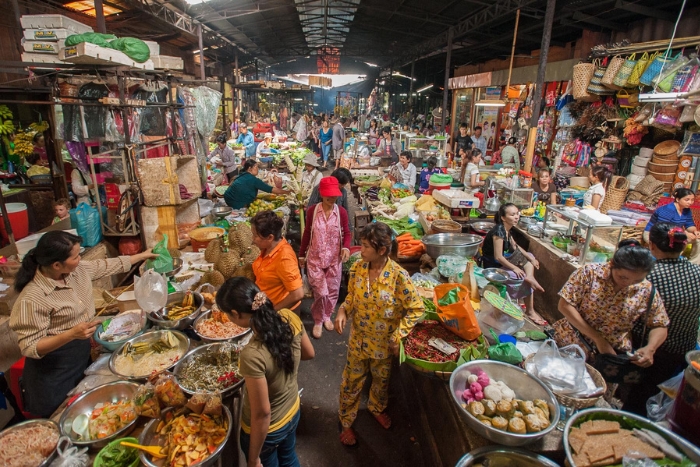 A bustling local market in Phnom Penh, Cambodia, filled with vibrant stalls selling fresh produce, cooked meals, and various goods, with people shopping and engaging in lively conversation.