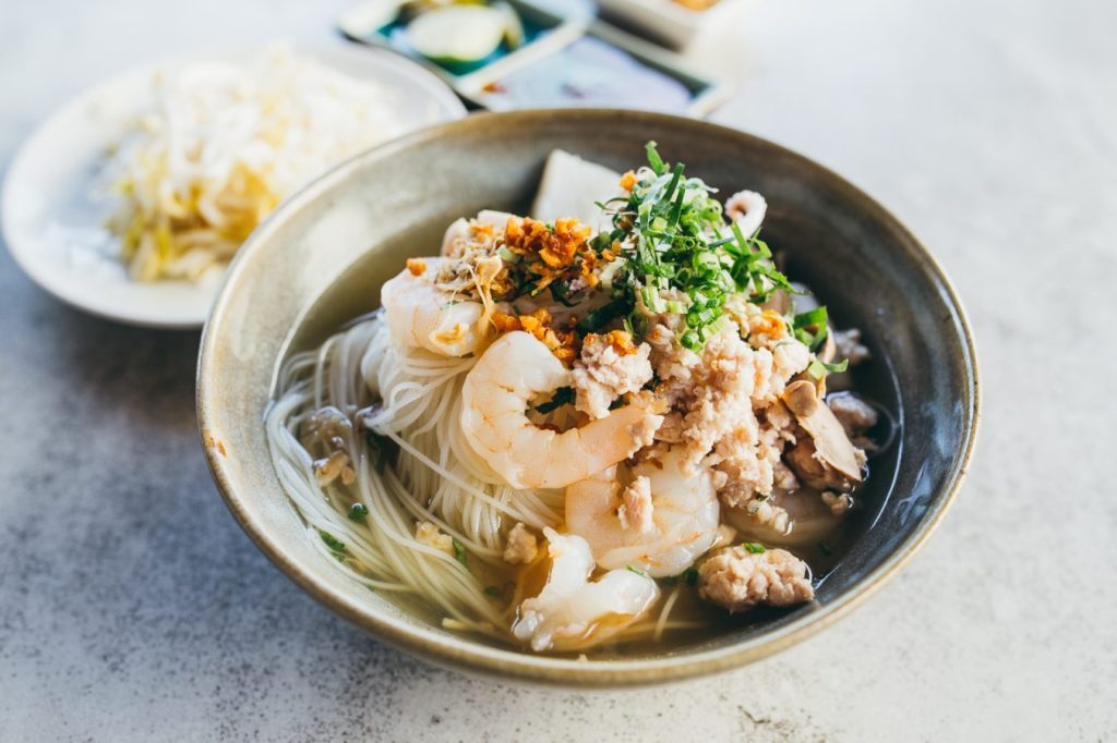 A bowl of Cambodian Kuy Teav noodle soup topped with shrimp, ground meat, and herbs, served alongside a plate of bean sprouts.