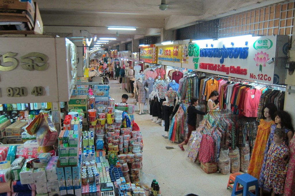 Interior of a market featuring various stalls with colorful clothing, snacks, and household items, along with shoppers browsing in Phnom Penh.