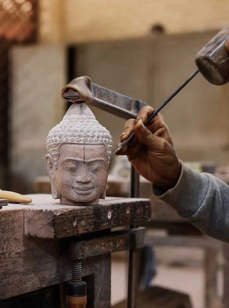 A close-up of a sculptor's hand carving a stone Buddha head on a workbench, showcasing the intricate details of the sculpture.