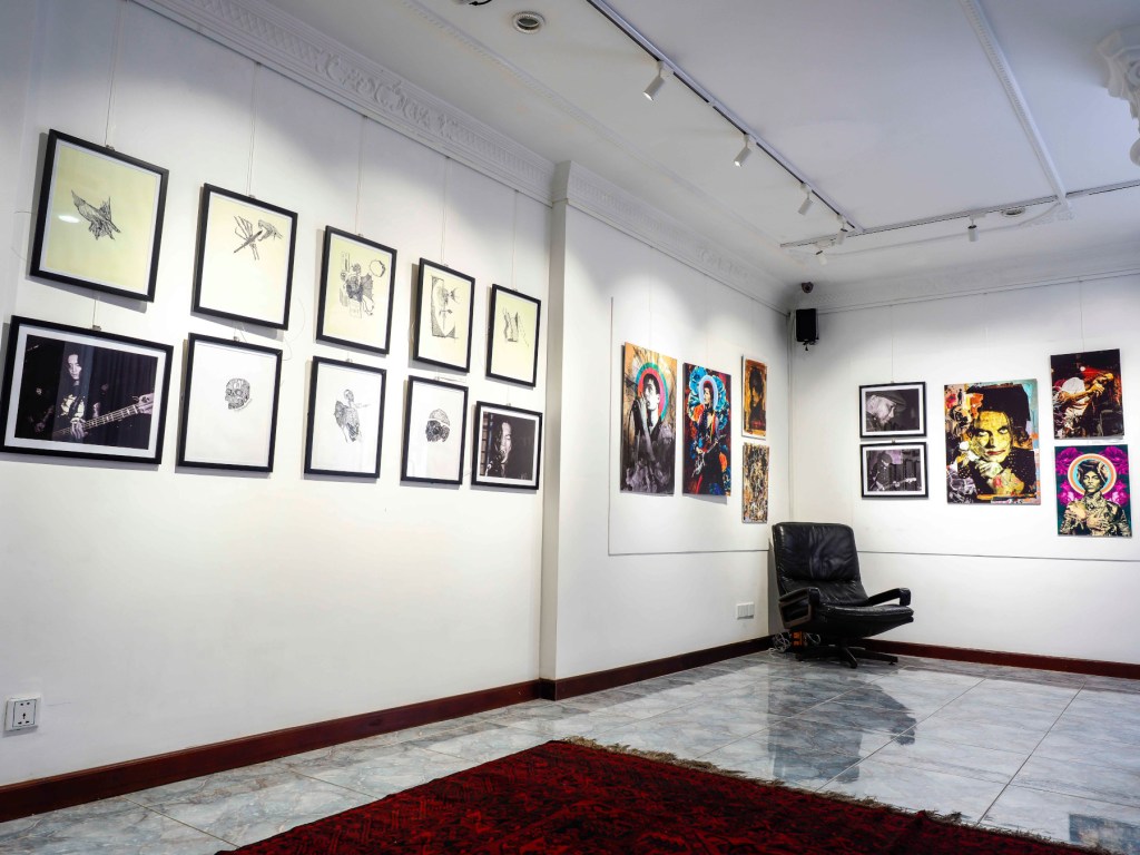 Art gallery interior featuring a collection of framed artworks and photographs on white walls, with a black chair and decorative carpet on the floor.