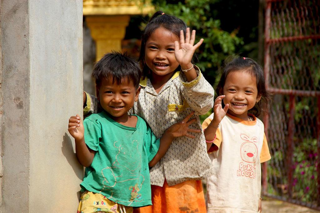 Three smiling children standing together, waving at the camera, in a rural setting.