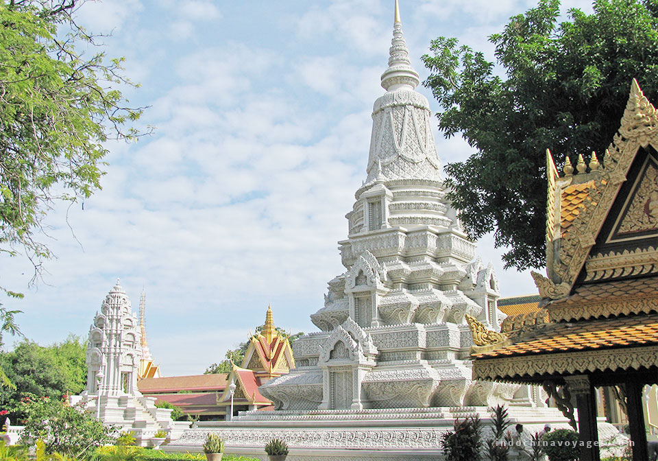 A white multi-tiered stupa surrounded by lush greenery, with traditional Cambodian roofs in the background under a partly cloudy sky.