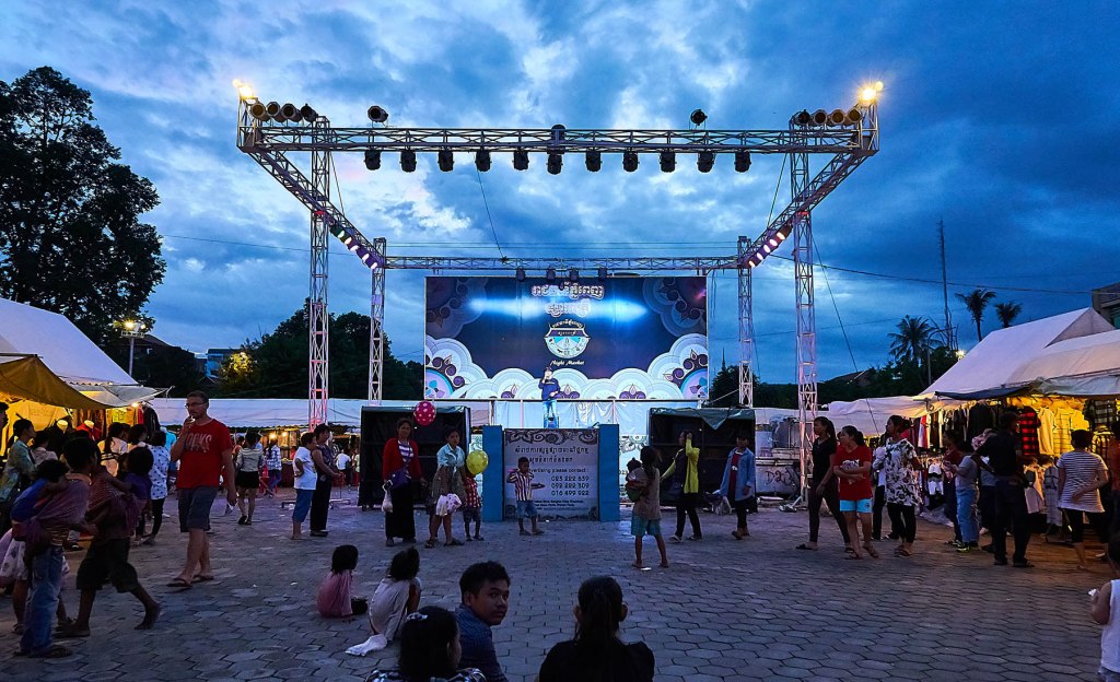 Evening market scene with a stage set up for performances, surrounded by tents and people walking around, under a cloudy sky.