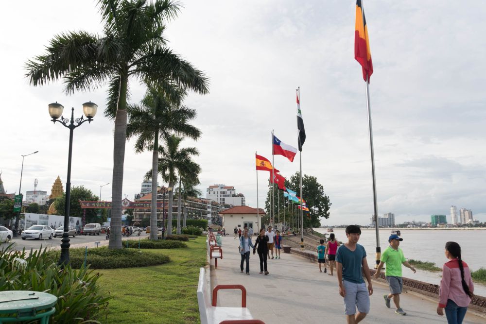 A vibrant walkway beside the river in Phnom Penh, Cambodia, with palm trees and people strolling. Flags from various countries line the pathway, and city buildings are visible in the background under a cloudy sky.