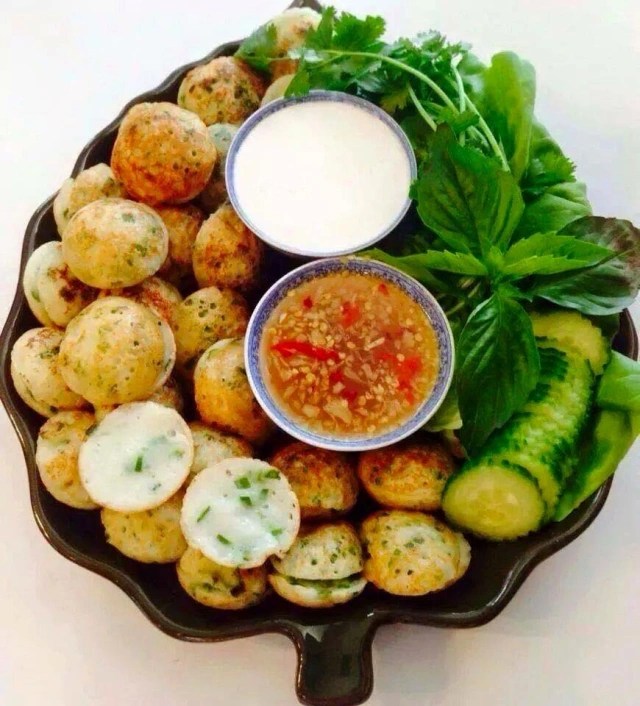 A platter of Num Kruk Khmer, featuring round coconut rice cakes topped with green onions, served with dipping sauces and fresh herbs, including cucumber and leafy greens.