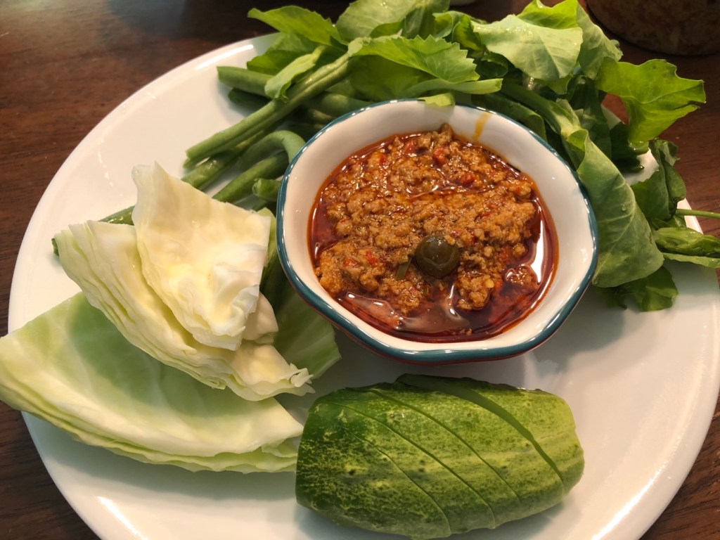 A plate featuring a bowl of Prahok Ktis (fermented fish dip) surrounded by fresh vegetables, including cabbage, cucumber, green beans, and leafy greens.