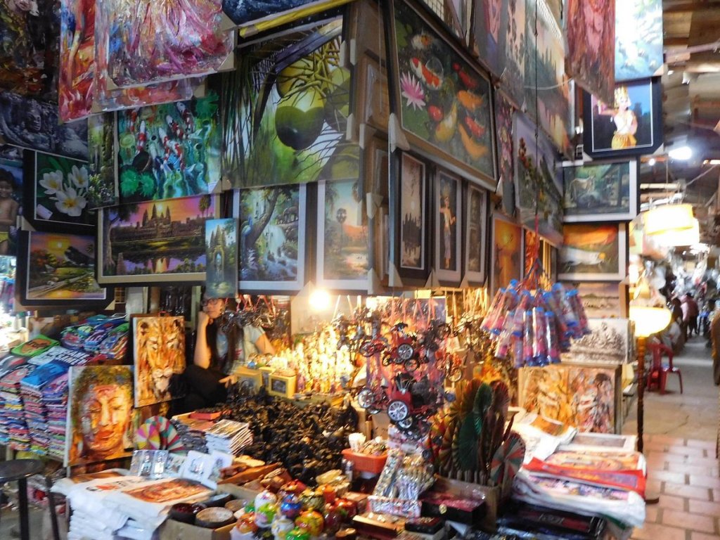 A vibrant market scene showcasing various art pieces and souvenirs, with colorful paintings hanging on the walls and a vendor seated among handcrafted items.