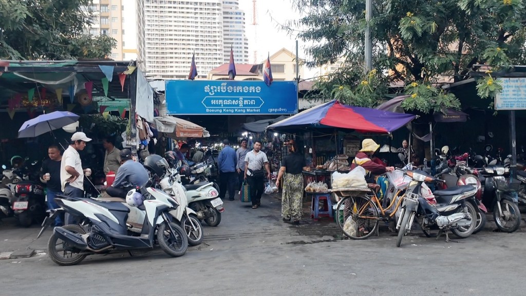 View of the bustling Boeung Keng Kang Market in Phnom Penh, with vendors under colorful awnings and shoppers navigating through parked motorbikes.