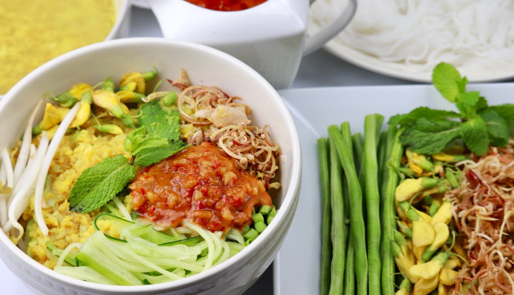 A bowl of authentic Khmer noodles topped with fresh herbs, shredded coconut, and a spicy chili sauce, accompanied by green vegetables and condiments.
