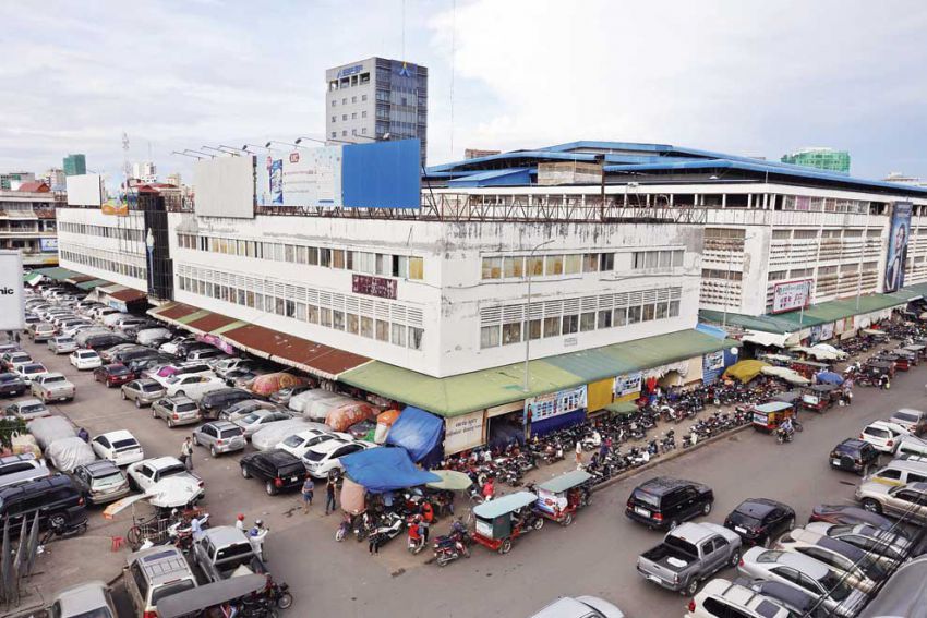 Aerial view of a busy market area in Phnom Penh, Cambodia, with vehicles parked alongside a large building and vendors setting up stalls.